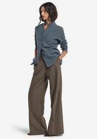 Kvinna iförd en strukturerad blå cardigan med knappar och vida bruna byxor, stående med händerna i fickorna mot en vit bakgrund.