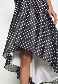 Kvinna iklädd en svart och vit prickig asymmetrisk kjol, med ett ben och en hand synlig mot en enfärgad bakgrund.
