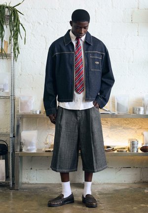 Muž v tmavom rifľovom saku, bielej košeli s červenou pruhovanou kravatou, širokých sivých šortkách, bielych ponožkách a tmavých mokasínach, stojaci v interiéri.