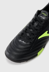 Sapatilha desportiva preta em couro com atacadores pretos, detalhes em amarelo neon e marca "Joma Aguila" na língua e na área do dedo do pé.