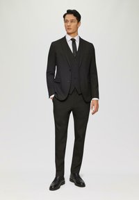 Schwarzer Anzug mit einem Sakko mit drei Knöpfen, passender Weste und maßgeschneiderten Hosen. Weißes Hemd und schwarze Krawatte. Glänzende schwarze Eleganzschuhe.
