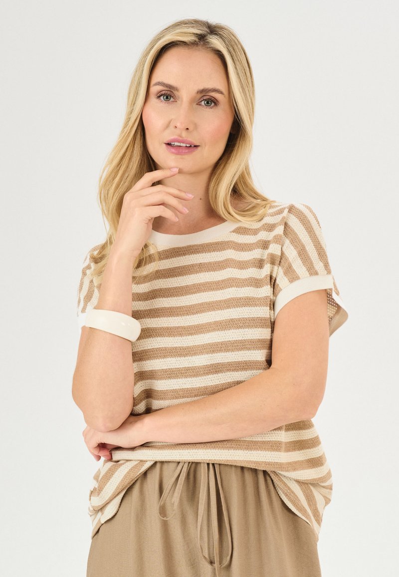 Beige en wit gestreept gebreid topje met korte mouwen, gecombineerd met een lichtbruine strikrok, en een glanzende witte armband om de arm.