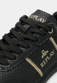 Black sneakers with a textured reptile-like surface, gold accents, flat black laces, and a prominent "REPLAY" logo on the side.