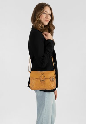 Bolso cruzado amarillo de cuero con textura suave, correa ajustable, cierre de hebilla y detalle de encanto de ancla, llevado al hombro sobre un blazer negro.