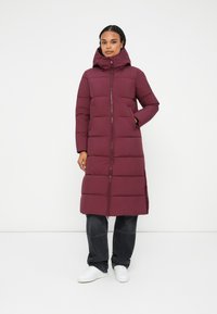 Geacă puffer bordo cu fermoar complet și glugă, având cusături orizontale, buzunare laterale și un design până la genunchi. Purtați-o cu pantaloni închiși la culoare și pantofi albi.