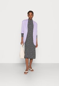 Blazer oversized lavanda sobre um vestido preto com padrão floral, combinado com sandálias brancas de salto e uma bolsa texturizada bege.