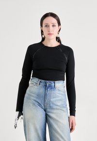 Schwarzes Langarmoberteil mit rundem Ausschnitt und dekorativen Metallteilen, kombiniert mit hellblauen, hochtaillierten Jeans aus Denim. Glatte Stofftextur.