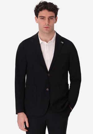 Uomo con blazer nero e camicia bianca con colletto, in piedi con una mano in tasca contro uno sfondo semplice.