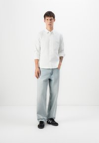 Joven de pie con las manos en los bolsillos, vistiendo una camisa blanca de botones, unos jeans de pierna ancha azul claro y zapatillas negras con rayas blancas.