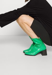 Vihreät tekokrokotiilinahasta valmistetut nilkkakengät, joissa on laskostettu muotoilu, neliönmuotoinen kärki ja tuhti musta korko, aseteltuna neutraalia taustaa vasten.