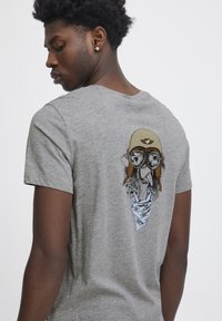 Grå bomulds t-shirt med et farverigt grafisk motiv af en karakter i goggles og pilot-hjelm på ryggen. Korte ærmer og afslappet pasform.