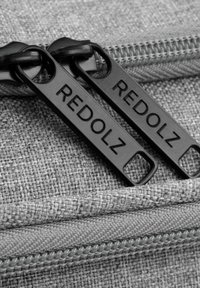 Dettaglio di una borsa di tessuto grigia con due zip nere etichettate "REDOLZ", realizzata in materiale testurizzato e con cuciture visibili lungo la cerniera.