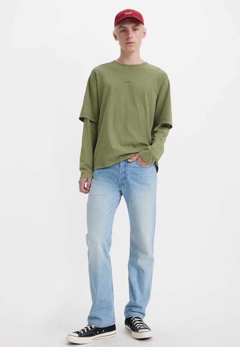 Chemise verte à manches longues avec une coupe décontractée, jean bleu clair et baskets en toile noires, surmontée d'une casquette bordeaux affichant un logo.