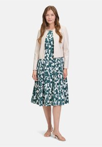 Femme debout, portant une robe midi à fleurs verte et blanche, une veste courte beige clair et des sandales à talons bas beiges avec des lanières.