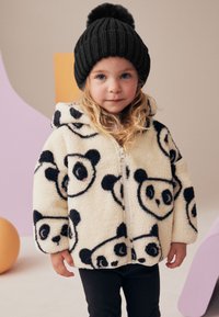 Cremefarbige, flauschige Jacke mit schwarzen Pandamustern, vorne mit Reißverschluss und Kapuze. Schwarze gestrickte Mütze mit Bommel oben.