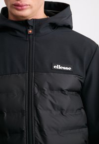 Černá péřová bunda s vysokým límcem a kapucí. Obsahuje zipový uzávěr, texturovanou spodní část a bílé logo "ellesse" na hrudi.