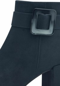 Bottine en daim bleu marine avec une large sangle et une boucle noire carrée. Caractérisée par des lignes épurées et une texture lisse.