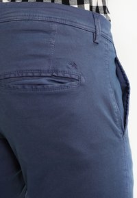 Mörkblå bomullsbyxor har en bakficka med klaff och ton-i-ton broderi. Texturen är slät med en lätt lyster.