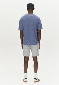 Blå t-shirt med kort ärm, grå rutiga shorts, vita strumpor och svarta sneakers sedd bakifrån mot en helt vit bakgrund.