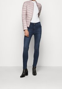 Chaqueta acolchada rosa claro, top blanco ajustado, jeans skinny azul oscuro y botas negras de tobillo con tacón grueso. Atuendo simple y moderno.