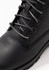 Bota de cuero negra con cordones grises, ojales de metal y superficie texturizada. Presenta acentos de costura y una suela robusta para mayor durabilidad.