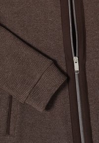 Brązowa żakardowa dzianinowa rękaw i przód kurtki z srebrnym zamkiem błyskawicznym i dopasowaną brązową lamówką, pokazująca szczegóły kieszeni.