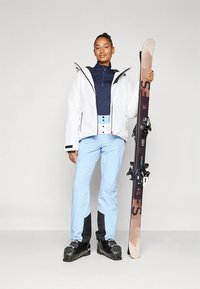 Costumul de schi include o geacă izolată albă cu accente negre, un tricou bleumarin, pantaloni bleu deschis și cizme de schi negre. Ține schiurile în mână.