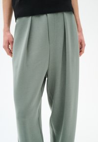 Pantaloni in tessuto verde chiaro con una texture liscia, caratterizzati da pieghe laterali e una silhouette rilassata. Fascia in vita visibile nella parte superiore.