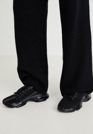 Schwarze gestrickte Hosen, die schwarze strukturierte Sneakers mit dicken Sohlen auf hellem Bodenhintergrund bedecken.