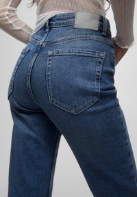 Jeans de mezclilla azul de tiro alto con un diseño de pierna recta, que cuentan con bolsillos traseros y un parche de marca gris claro en la cintura.