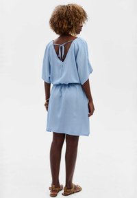 Jasnoniebieska sukienka z miękkiego materiału, charakteryzująca się luźnym krojem, krótkimi rękawami, otwartym tyłem z wiązaniem oraz marszczoną talią.