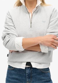 Sudadera con cremallera de color gris claro y cuello alto, con puños acanalados y un pequeño logotipo en el pecho, superpuesta sobre una camisa blanca.