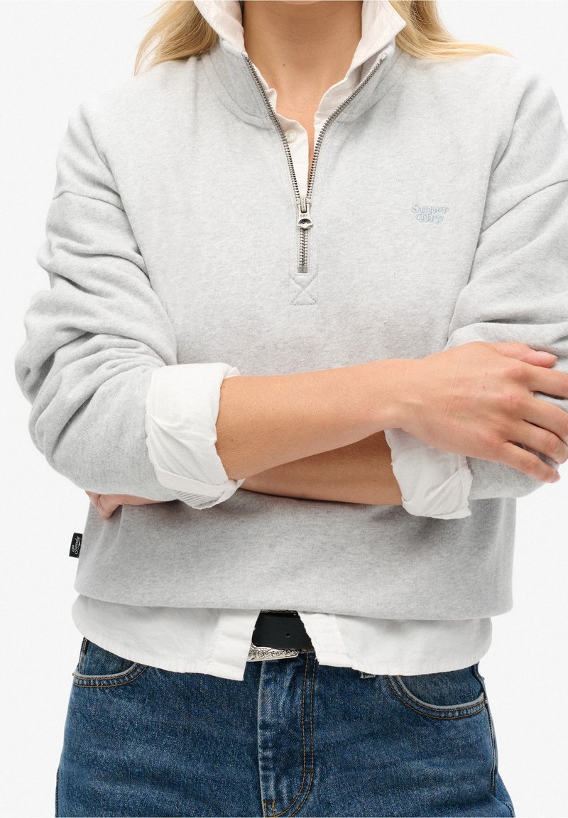 Sudadera con cremallera de color gris claro y cuello alto, con puños acanalados y un pequeño logotipo en el pecho, superpuesta sobre una camisa blanca.