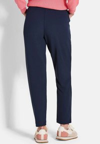 Personne portant un pantalon slim-fit bleu marine, un pull côtelé corail et des baskets beiges avec des accents roses, se tenant contre un fond blanc.