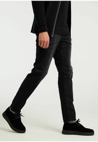 Zapatillas negras con material texturizado, que cuentan con cordones y una suela gruesa, combinadas con jeans negros ajustados. El atuendo es llevado por una persona que camina.