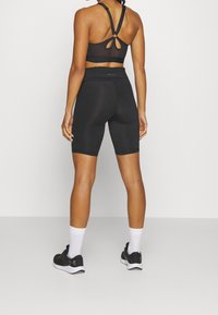 Czarny sportowy stanik z siateczkowym tyłem, zestawiony z czarnymi szortami z wysokim stanem. Modelka nosi czarne sneakersy z białymi podeszwami oraz skarpetki do kostek.