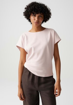 Donna con capelli ricci che indossa una maglietta rosa chiaro a maniche corte e pantaloni marrone scuro, in piedi davanti a uno sfondo neutro.