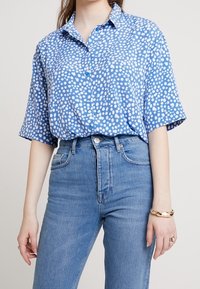 Ljusblå kortärmad skjorta med ett vitt prickigt mönster, med knappad krage, i kombination med högmidjade blå jeans. Slät tygkvalitet.