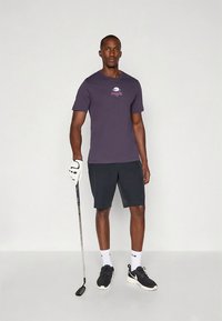 Vīrietis tumšā Nike T-kreklā, melnā šortos un sporta apavos, turot golfa nūju un stāvot uz vienkārša gaiša fona.