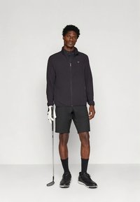 Casaco atlético preto com fecho de correr frontal, combinado com calções pretos. Modelo segura um taco de golfe e usa luvas brancas de golfe e sapatos pretos.