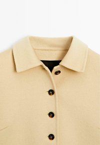 Beige wollen blendjas met een kraag, vijf donkere knopen en een zachte textuur. Inclusief twee zijzakken met minimale stiksels.