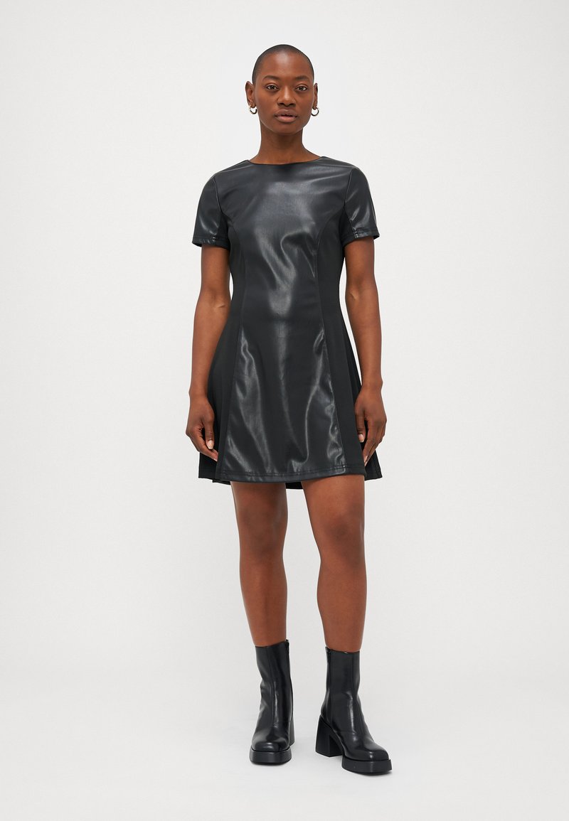 Schwarzes Kurzkleid aus Kunstleder mit rundem Ausschnitt, kurzen Ärmeln und einem ausgestellten Schnitt. Kombiniert mit kniehohen schwarzen Plateaustiefeln.
