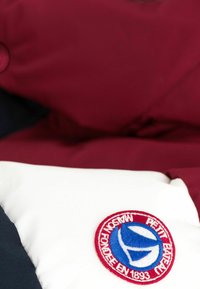 Veste rembourrée colorée avec une partie supérieure bordeaux, une partie inférieure bleu marine foncé et un patch blanc avec un logo brodé en bleu et rouge.