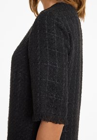 Schwarzes strukturiertes Kleid mit kurzen Ärmeln. Der Stoff weist ein gewebtes Muster und fransige Ärmelenden auf. Minimalistisches Design, keine sichtbaren Verschlüsse.