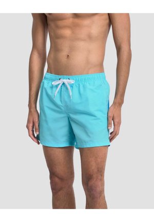 Mann trägt hellblaue Badeshorts mit weißem Kordelzug und steht vor einem schlichten hellgrauen Hintergrund.