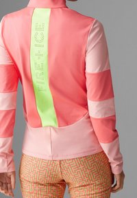 Rosafärgad träningsjacka med långa ärmar, med en neongrön bakpanel och varumärket i perforerad detalj. Flera nyanser av färgblock.