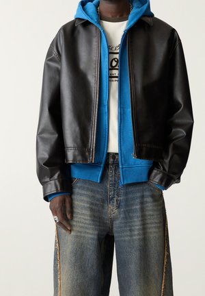Personne portant une veste en cuir noire sur un hoodie bleu zippé, un t-shirt blanc avec un graphisme, un jean bleu délavé avec des coutures latérales effilochées, et des bagues en argent.