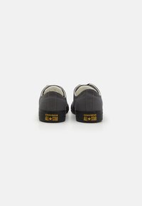 Sapatilhas de canvas cinza com solas de borracha preta. Apresentam um design de cano baixo e o logótipo amarelo "CONVERSE ALL STAR" no calcanhar. Sem atacadores visíveis.