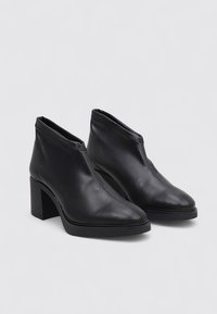 Une paire de bottines en cuir noir avec des talons blocs, des bouts pointus et un minimum de coutures sur un fond clair.