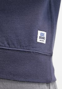 Granatowy sweter wykonany z dzianiny bawełnianej, z małą białą metką z czarnym i niebieskim tekstem blisko dolnego brzegu. Gładka faktura.